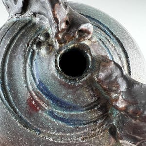 Puede incluir: Primer plano de un recipiente de cerámica hecho a mano con una superficie oscura y texturizada. El recipiente presenta un diseño circular con crestas concéntricas y una abertura oscura. Acentos de azul y rojo añaden color.