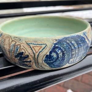 May include: A handcrafted ceramic bowl with a light green interior and a textured exterior. The bowl features a decorative pattern in shades of blue and brown, adding a unique artistic touch to the piece. This bowl is perfect for serving or display.