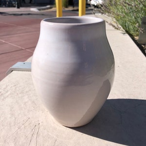May include: A white ceramic vase with a rounded body and a wide opening. The vase is sitting on a light gray surface.