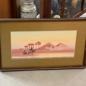 Puede incluir: Una pintura enmarcada que representa un paisaje desértico con una caravana de camellos viajando por las dunas de arena. La pintura presenta una paleta de colores cálidos con tonos de naranja, amarillo y marrón. La caravana de camellos está silueteada contra el sol poniente, creando un efecto dramático.