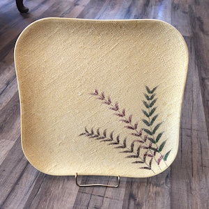 Puede incluir: Una bandeja cuadrada de servir de color crema con textura tejida y un diseño de helecho en tonos verdes y morados.