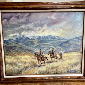 May include: Oil painting depicting a landscape with three figures on horseback, traversing a field towards a snow-capped mountain range. The artwork is framed in a dark wood frame with a white inner border. The color palette includes earth tones and blues.