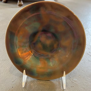 Puede incluir: Un cuenco de cerámica con un patrón de remolino de esmalte marrón, verde y azul. El cuenco está sobre un soporte de acrílico transparente.