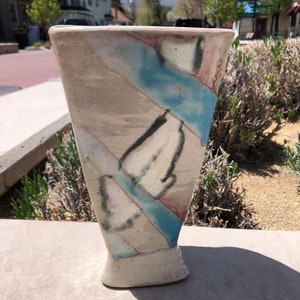 May include: A white ceramic vase with a blue, pink, and gray abstract design. The vase has a wide mouth and a narrow base.