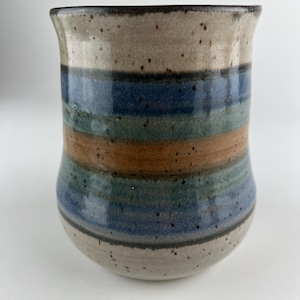 May include: A handmade ceramic mug with a speckled, light beige top and bottom. The mug features horizontal bands of blue, green, and brown. The mug has a slightly flared rim and a rounded base. The mug is suitable for hot or cold beverages.