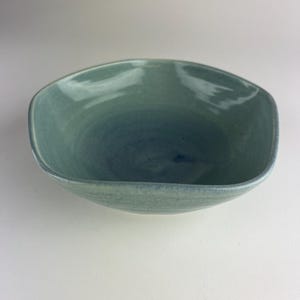 Könnte beinhalten: Eine handgefertigte, quadratisch geformte Keramikschale mit hellblauer und türkisfarbener Glasur. Die Schale hat eine glatte, glänzende Oberfläche und eine leicht unregelmäßige Form, was darauf hindeutet, dass sie handgefertigt ist. Das Innere hat einen dunkleren Blauton.