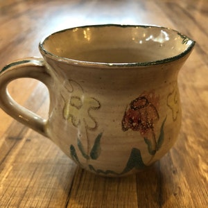 Könnte beinhalten: Eine kleine, cremefarbene Keramiktasse mit Henkel. Die Tasse hat einen grünen Rand und ist mit handbemalten Blumen in Gelb-, Orange- und Grüntönen verziert.