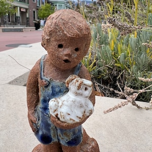 May include: A ceramic sculpture of a child holding a small white animal. The child is wearing blue overalls and has a brown, textured surface. The sculpture is on a brown base.