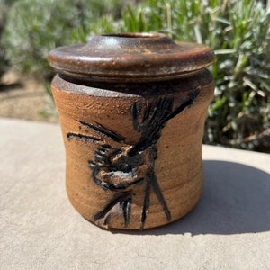 Può includere: Un piccolo vaso di ceramica marrone con un coperchio. Il vaso ha un disegno nero di un uccello su un ramo.