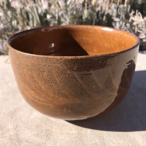 Puede incluir: Un tazón de cerámica marrón con un esmalte moteado. El tazón es redondo y tiene un ligero borde.