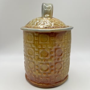 Puede incluir: Un tarro cilíndrico de cerámica con tapa a juego que presenta un asa vertical. La superficie exterior está esmaltada en tonos cálidos de naranja, amarillo y marrón, decorada con un patrón geométrico en relieve de cuadrados y círculos.