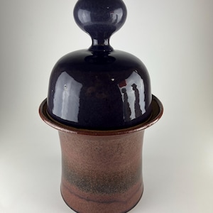 May include: A ceramic jar with a dark purple lid and a round handle. The jar's body is a reddish-brown color with darker bands near the base. The lid sits on a slightly flared rim. The jar has a glossy finish.