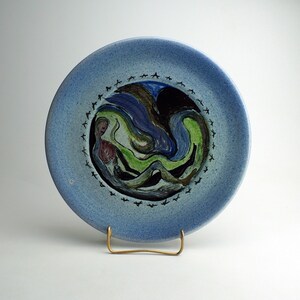 May include: A blue ceramic plate with a painted design of a swirling abstract pattern in shades of green, blue, black, and brown. The plate has a decorative border of black birds.