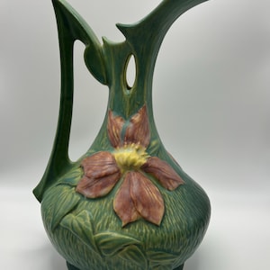 Puede incluir: Jarra de cerámica verde con un gran diseño floral estilizado en tonos rojos y amarillos. La jarra tiene un asa y un pico únicos, y la flor está en relieve en la parte delantera.