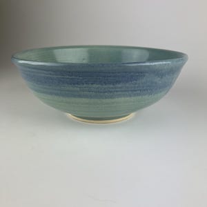 Könnte beinhalten: Eine handgefertigte Keramikschale mit blauer und grüner Glasur. Die Schale hat eine breite, flache Form und eine glatte, glänzende Oberfläche. Das Äußere weist horizontale Farbstreifen auf, die von Grün nach Blau übergehen. Der Boden ist hellbeige.