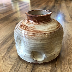 Könnte beinhalten: Eine weiße Keramikvase mit brauner Glasur und einer strukturierten Oberfläche. Die Vase hat eine abgerundete Form mit einer breiten Öffnung oben.