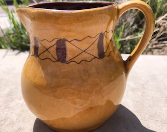 May include: A yellow ceramic pitcher with a brown handle and brown geometric designs. The pitcher has a rounded body and a wide mouth.