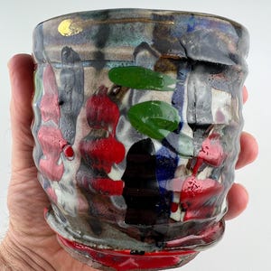 Op de afbeelding: Een handgemaakte keramische beker met een getextureerd oppervlak. De beker heeft een mix van kleuren, waaronder rood, groen, zwart en blauw, met gouden accenten. Het ontwerp is abstract en artistiek, voor dranken.