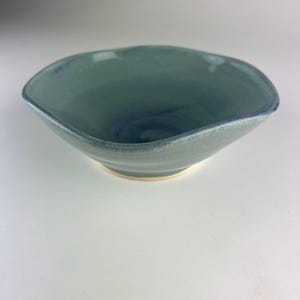 Könnte beinhalten: Eine weiße Keramik-Tasse mit schwarzem Henkel und dem Text "But First, Coffee" in fetter, schwarzer Schrift. Die Tasse steht vor einem verschwommenen Hintergrund, der eine gemütliche Umgebung suggeriert.