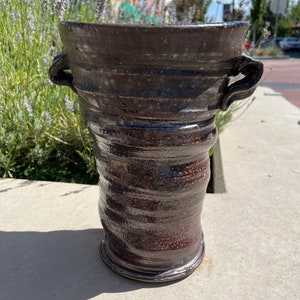 Könnte beinhalten: Eine dunkelbraune Keramikvase mit zwei Henkeln. Die Vase hat ein strukturiertes, geschichtetes Design und eine glänzende Oberfläche.