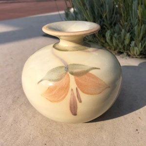 Puede incluir: Un pequeño jarrón de cerámica de color crema con cuerpo redondeado y cuello estrecho. El jarrón está decorado con un diseño pintado a mano de flores estilizadas en tonos marrones, naranjas y verdes.