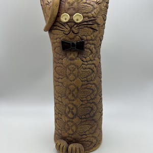 May include: A tall, cylindrical ceramic cat sculpture in a tan, textured finish with a repeating floral pattern. The cat features a small, dark brown bow tie, two round white eyes, a curved hat on one ear, and two small paws at the base.