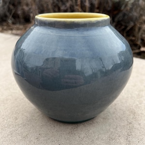 May include: A round, ceramic vase with a glossy, blue-gray exterior and a yellow interior rim. The vase has a smooth, rounded shape and sits on a concrete surface. The background features blurred greenery.