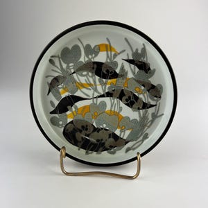 Può includere: Un piatto decorativo con bordo nero e un motivo floreale nei toni del grigio, nero e giallo. Il piatto è esposto su un supporto in metallo color oro. Il design presenta fiori e foglie stilizzati.