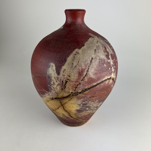 May include: A handcrafted, red ceramic vase with a unique, organic shape. The vase features a textured surface with beige and brown accents, and a narrow neck. The design includes a network of dark lines, adding to its artistic appeal.