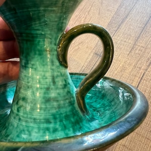 Op de afbeelding: Een groene keramische olielamp met een handvat en een brede basis. De lamp heeft een geglazuurde afwerking en een rustieke uitstraling.