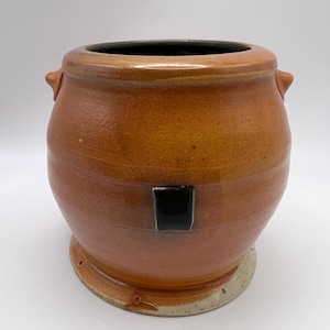 May include: A handcrafted, brown ceramic pot with a black interior and a black square accent. The pot has two small handles and sits on a circular base. The exterior has a textured, earthy appearance.