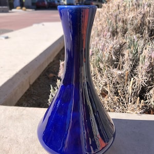 May include: A cobalt blue ceramic vase with a rounded, bulbous body and a narrow neck. The vase has a glossy finish and reflects light.