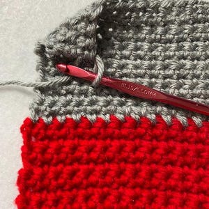 May include: Close-up of crochet work in progress. The project features alternating rows of red and gray yarn. A red crochet hook is inserted into the gray yarn, with the hook's size marking visible. The texture of the yarn is clearly visible.