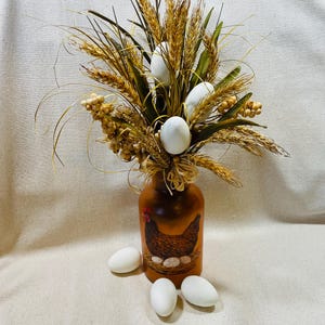 May include: A decorative arrangement featuring a brown ceramic vase with a hen and egg design. The vase holds a bouquet of wheat, greenery, and white eggs. Additional white eggs are scattered around the base, creating a rustic, farmhouse aesthetic.