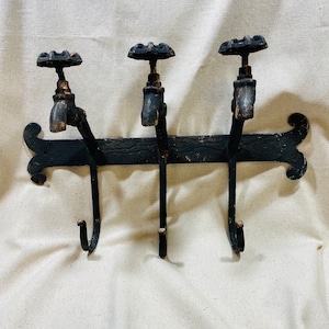 May include: Black metal coat rack with three hooks shaped like antique faucets. The hooks are attached to a black metal bar with decorative scrollwork on the ends.
