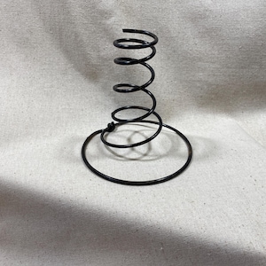 May include: Black metal spring-shaped vase. The vase has a circular base and a spiral design that rises upwards. The vase is made of thin metal and has a matte finish. The vase is designed to hold flowers or other decorative items.