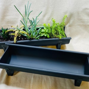May include: Two black metal planters, one with a variety of small plants, the other empty. The planters are shaped like gutters and have metal brackets for hanging.