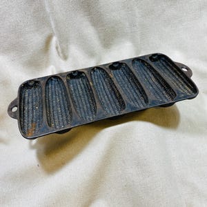 May include: A dark, rectangular cast iron corn stick pan featuring seven individual, elongated, ear-of-corn-shaped molds. The metal surface shows a textured, dark grey and black finish with small areas of rust visible in the bottom of the molds.