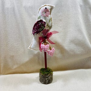 May include: A decorative bird figurine with a white body, embroidered pink flowers, and a burgundy wing. It has a red stem with pink gingham hearts and a moss-covered wooden base. A pink ribbon is tied around the stem.