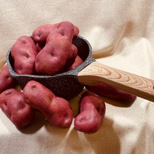 May include: A small black pot with a wooden handle filled with red potatoes. The pot is sitting on a beige fabric surface.