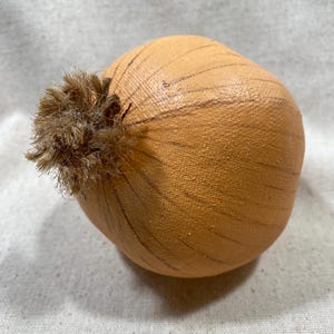 May include: A decorative, handcrafted onion with a light brown hue and textured surface. The onion features subtle, dark brown lines and a tuft of brown fibers at the top, resembling the onion's stem. This item is likely a decorative piece for a kitchen or dining area.