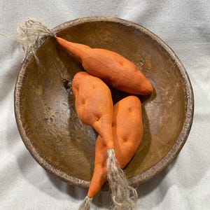 May include: Three orange fabric carrots with burlap stems in a brown wooden bowl.