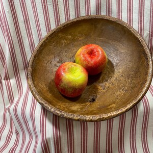 May include: A rustic wooden bowl holds two apples. The apples are red and yellow, with a slight green hue. The bowl is a warm brown color and has a rough, textured surface. The background is a striped fabric with red and white stripes.