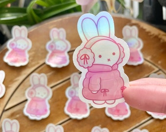 May include: A set of pink bunny stickers with iridescent accents. The bunnies are wearing pink winter coats and have white fur. The stickers are arranged on a wooden surface.
