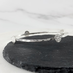 May include: A silver bangle bracelet with a hammered texture and four circular accents. The bracelet is displayed on a black surface, showcasing its simple yet elegant design. The bracelet is a piece of jewelry.
