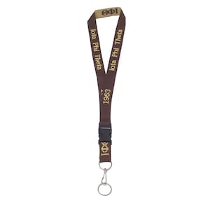 May include: A brown lanyard with gold text that reads "Iota Phi Theta" and "1963". The lanyard has a black plastic clasp and a metal ring at the bottom. The top portion of the lanyard is tan with the Greek letters "Iota Phi".