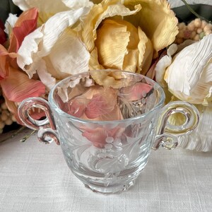 May include: A clear glass cup with a floral design and two handles. The cup is sitting on a white tablecloth.