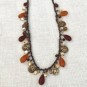 May include: A brown necklace with various decorative elements. The necklace features orange teardrop-shaped beads, small white beads, and gold-colored circular charms. The chain is a dark brown color, and the necklace is displayed on a white surface.