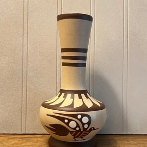 May include: A small, beige ceramic vase with a brown bird design. The vase has a narrow neck and a wide, rounded base.