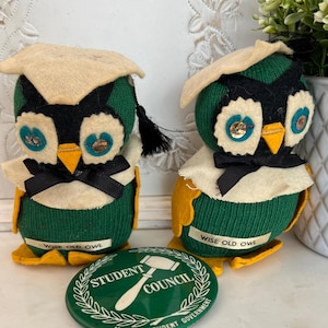 May include: Two vintage owl figurines wearing graduation caps, black bow ties, and yellow feet. The owls are green, black, and cream-colored. A green button with the text "Student Council" is in front of the owls. The owls have the text "Wise Old Owl" on their base.